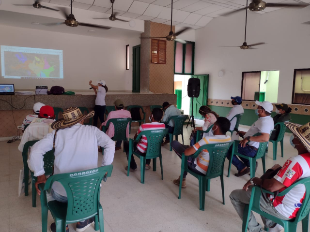 Fotografía de la socialización con la comunidad de San Benito Abad, Sucre, sobre el proceso de deslinde. En ella se ve a una mujer funcionaria de la ANT realizando la exposición y de espaldas sentados varios campesinos.