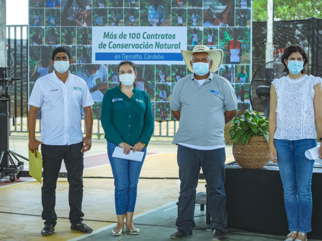 La Directora de la ANT, Myriam Martínez Cárdenas, aparece de pie y cubriendo sus bocas con tapabocas, con tres personas más, campesinos. De fondo una pancarta con varias fotografías de campesinos beneficiados de la entrega de títulos.