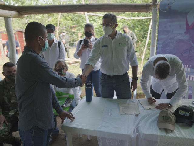 Agencia Nacional de Tierras entrega predios para la reincorporación efectiva de ex combatientes. En la fotografía se evidencian a cinco funcionarios de la ANT  vestidos de camisa blanca y con tapabocas, uno de ellos saludando de mano a un beneficiario de la entrega de predio. También se encuentra un soldado sentado al costado izquierdo