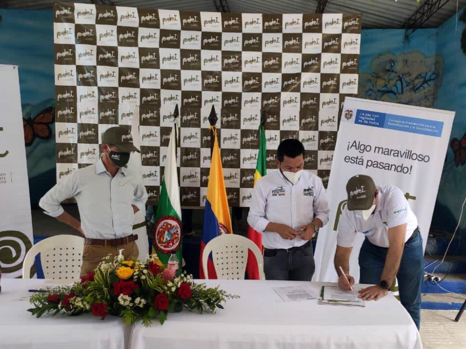 Tres hombres del equipo de la ANT en mesa de conversación, cada uno con tapabocas y camisa blanca, uno de ellos firmando un documento sobre la mesa. Encima de la mesa un arreglo de flores.