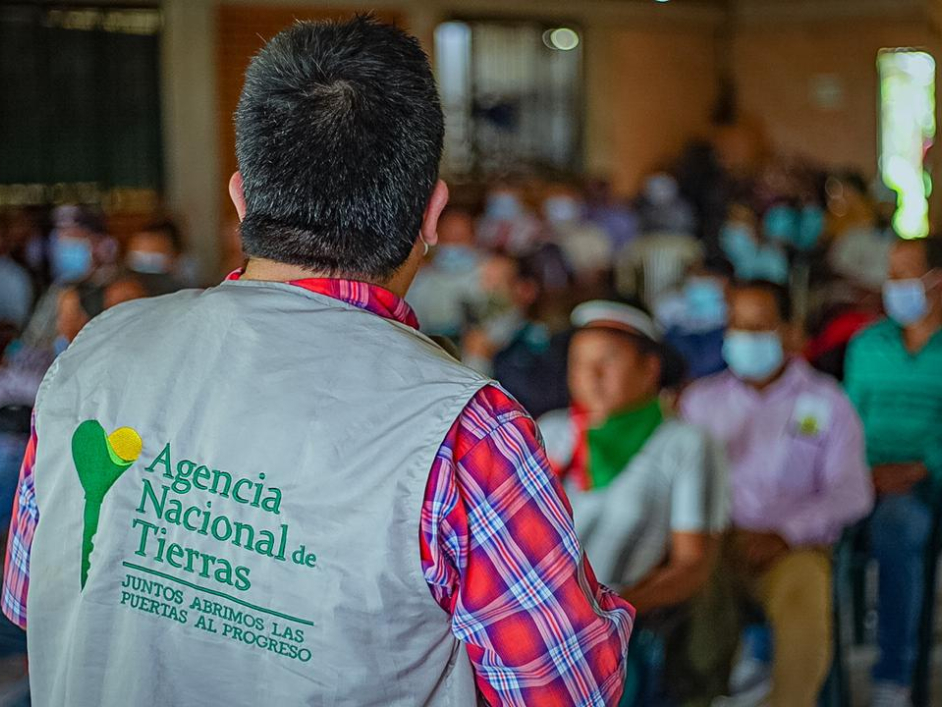 Fotografía de Audiencia Pública para la creación de la Zona de Reserva Campesina – ZRC de Santa Rosa, en Cauca. En ella aparece dando la espalda un funcionario de la ANT con su chaleco corporativo, hablando frente a varios campesinos quienes están sentados en sillas de plástico color azul.