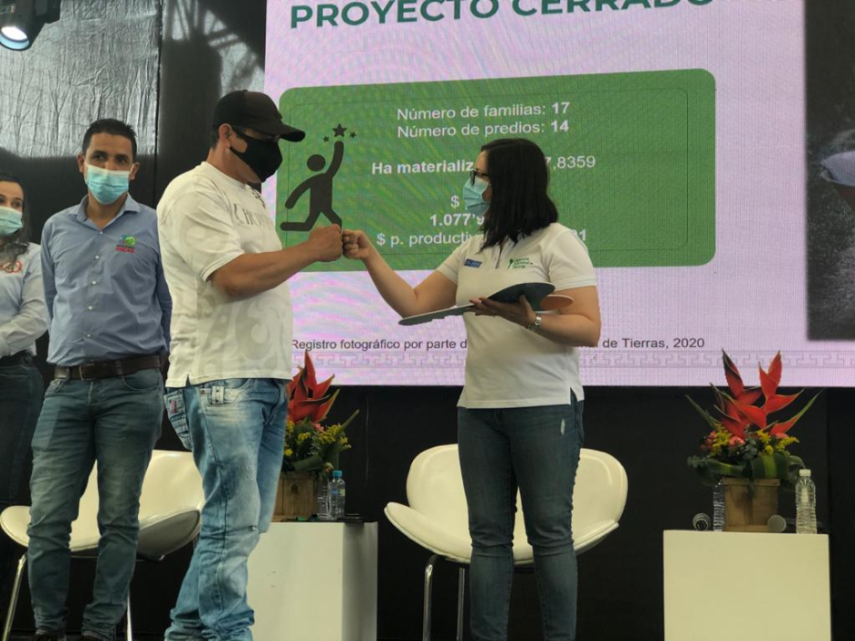Funcionaria de la ANT, vestida con camiseta blanca, pantalón azul y tapabocas, se da la mano con un hombre beneficiario. De fondo una pancarta informativa de la agencia, sillas blancas y dos personas más, un hombre y una mujer como espectadores.
